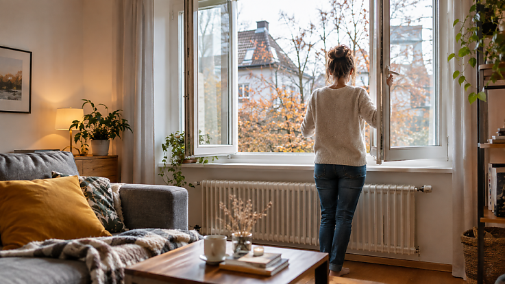 Fotorealistisches Wohnzimmer an einem Herbsttag. Eine Frau steht vor einem weit geöffneten Fenster und lüftet den Raum. Unter dem Fenster ist ein Heizkörper zu sehen. Im Wohnzimmer stehen ein Sofa, ein Couchtisch, Pflanzen und ein Regal. Durch das Fenster sind herbstlich verfärbte Bäume und Häuser zu sehen. Fotorealistisches Wohnzimmer an einem Herbsttag. Eine Frau steht vor einem weit geöffneten Fenster und lüftet den Raum. Unter dem Fenster ist ein Heizkörper zu sehen. Im Wohnzimmer stehen ein Sofa, ein Couchtisch, Pflanzen und ein Regal. Durch das Fenster sind herbstlich verfärbte Bäume und Häuser zu sehen.